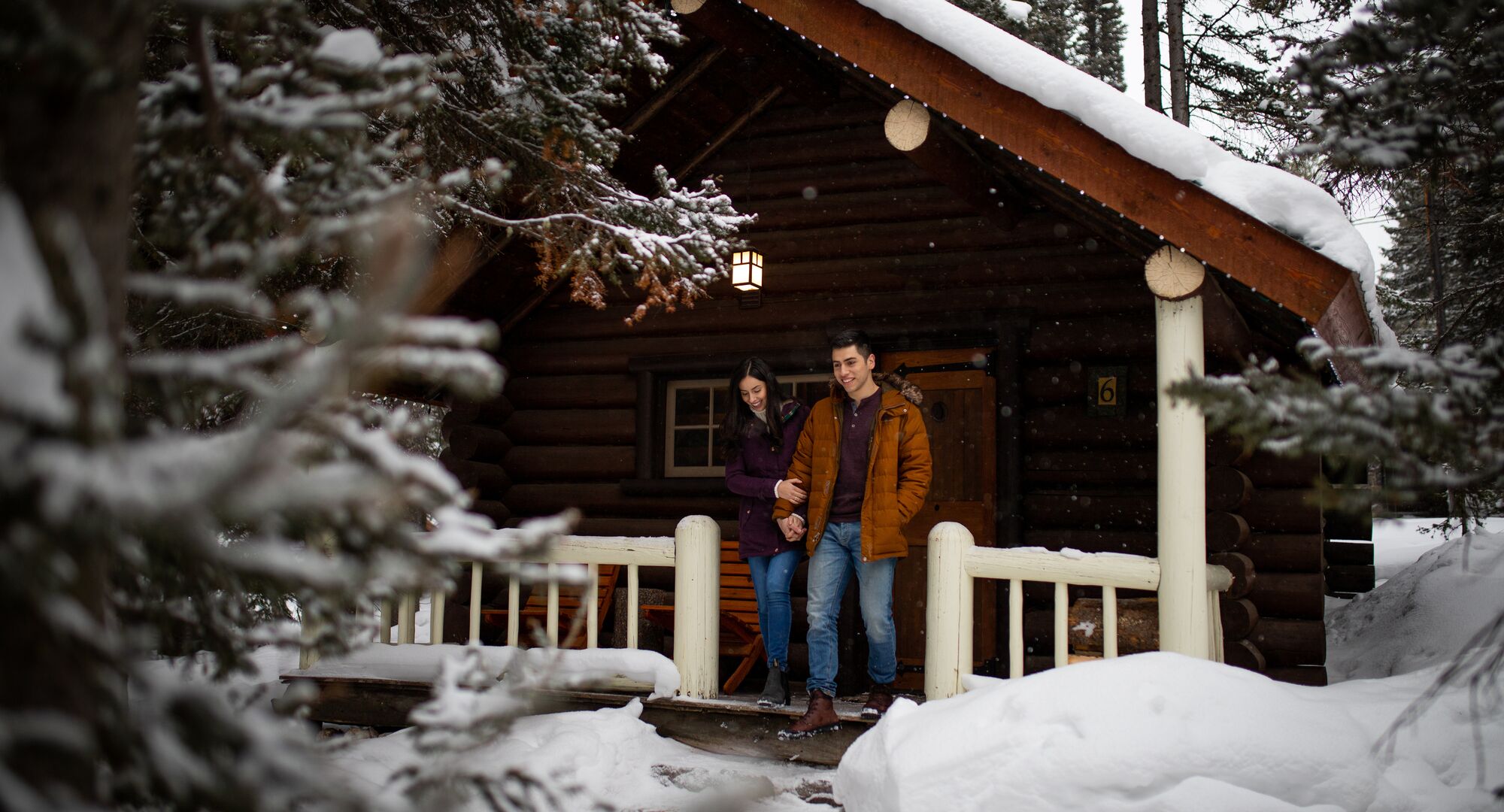 Two people on the front deck of a cabin nestled in a snow covered wooded area